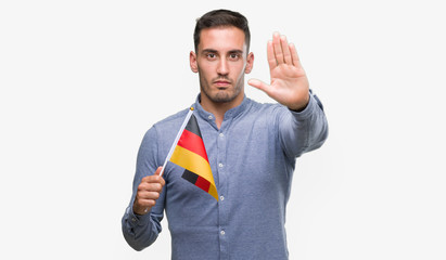 Sticker - Handsome young man holding a flag of Germany with open hand doing stop sign with serious and confident expression, defense gesture