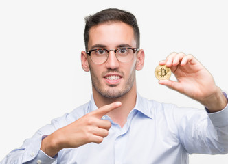 Sticker - Handsome young man holding bitcoin very happy pointing with hand and finger