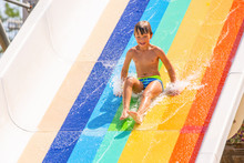 A Happy Boy On Water Slide In A Swimming Pool Having Fun During Summer Vacation In A Beautiful Aqua Park. A Boy Slithering Down The Water Slide And Making Splashes.