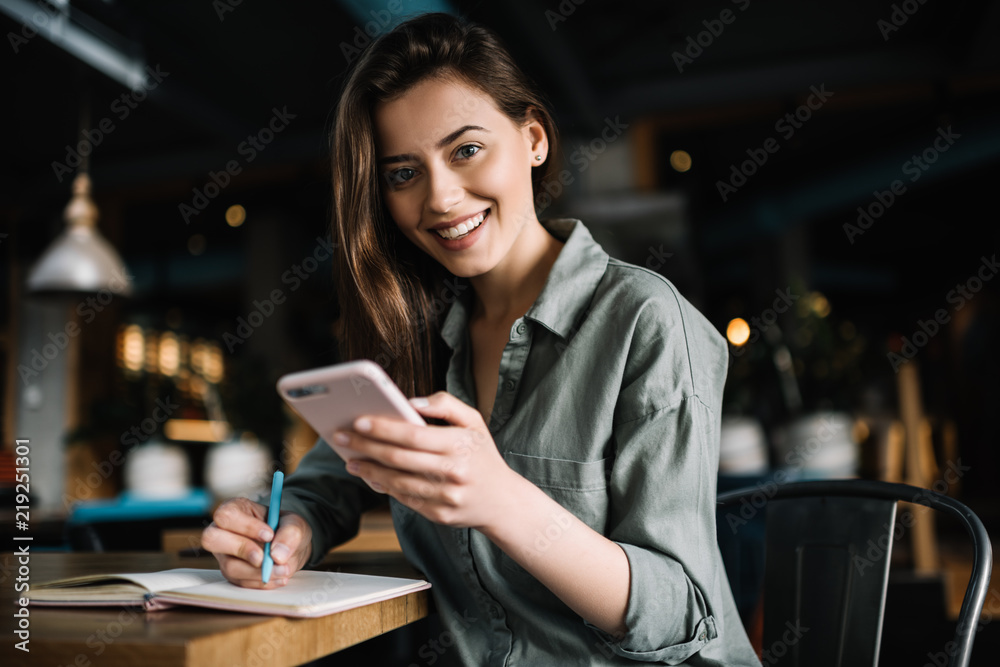Cheerful female using mobile phone for online banking, shopping web, learning  language sitting in loft coffee shop.   Positive caucasian woman holding modern smartphone  and making notes to study.  - obrazy, fototapety, plakaty 
