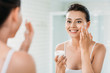 © LIGHTFIELD STUDIOS - beautiful happy girl applying face cream and looking at mirror in bathroom
