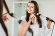 © LIGHTFIELD STUDIOS - happy brunette girl in bathrobe using hair curler at mirror in bathroom