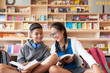 Teenage boy and girl sitting in library holding books and smiling on bookshelves background