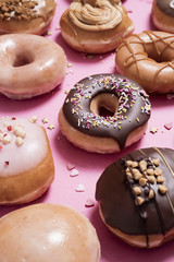 Closeup of classic chocolate ring donut (doughnut) on a pastel pink background with other donuts and sprinkles scattered around it