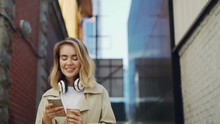 Slow Motion Of Happy Girl Using Smartphone While Walking In The Street Wearing Coat And Holding Take-away Coffee. Urban Lifestyle, Millennials And Young People Concept.
