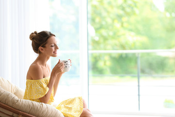 Sticker - Young woman with cup of aromatic coffee sitting in papasan chair near window at home