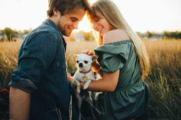 A young couple are sitting on a yellow autumn grass and hugging their white chihuahua breed dog.