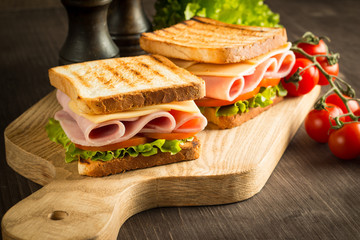 Close-up of two sandwiches with bacon, salami, prosciutto and fresh vegetables on rustic wooden cutting board. Club sandwich concept.