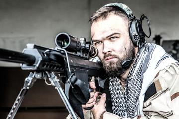 Portrait of U.S. Navy SEAL sniper on firing position, armed with large caliber sniper rifle with telescopic sight, wearing tactical headset with microphone, observing territory, searching targets