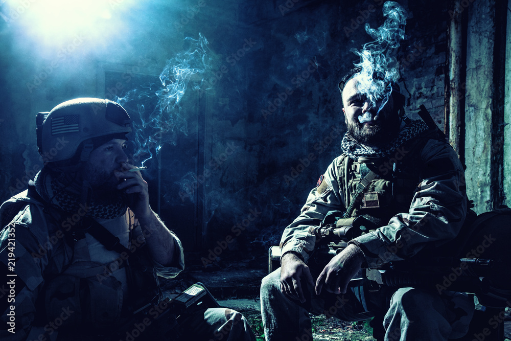Tired after hard battle army soldiers, exhausted with fight Navy Seal  rifleman sitting with assault rifle on knees, resting and smoking cigarette  in abandoned building, low key, high contrast shoot Stock-foto |