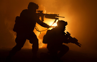 Silhouettes of two army soldiers, U.S. marines team in action, surrounded fire and smoke, shooting with assault rifle and machine gun, attacking enemy with suppressive gunfire during offensive mission