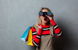 © Masson - Portrait of a young style blonde girl in red striped sweater with binoculars and shopping bags on grey background.