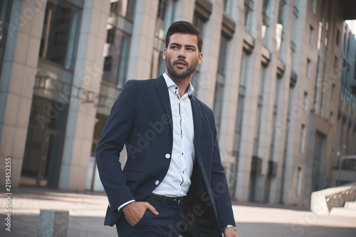 Plakat Confident businessman. Confident young man in full suit adjusting his sleeve and looking away while standing outdoors with cityscape in the background