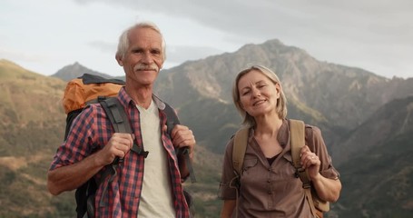 Wall Mural - Positive old caucasian couple trekking in mountains with backpacks, enjoying their adventure - portrait shot 4k