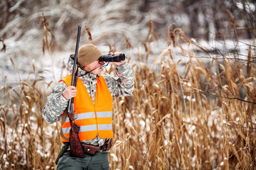 Male hunter in camouflage, armed with a rifle, standing in a snowy winter forest with duck prey