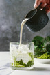 Cream pouring from jug to matcha green tea iced latte or cocktail in glass, with ice cubes, matcha powder on white marble table, decorated by green branches. Grey wall at background
