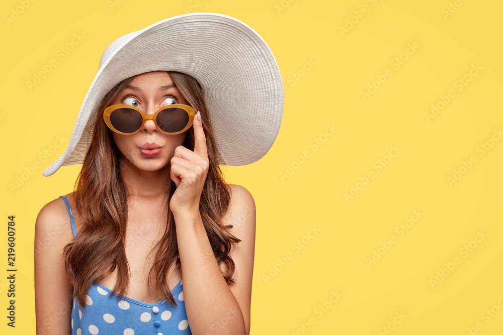 Stylish woman with surprised mysterious expression, looks asdie, wears stylish summer shades, hat and polka dot blouse, isolated over yellow background with copy space for your text. Relaxation - obrazy, fototapety, plakaty 