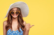© wayhome.studio  - Summer time concept. Cheerful beautiful brunette woman in stylish sunglasses and hat, has friendly smile, points with thumb aside, shows blank space for your advertisement or promotional text