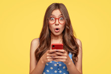 Surprised Young Female Model Holds Modern Cell Phone, Reads Sudden News On Internet Web Page, Surfes In Social Networks, Poses Against Yellow Background. People, Emotions, Technology, Lifestyle