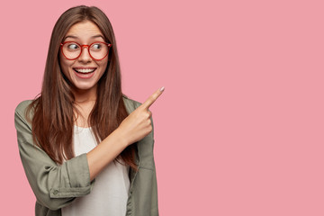 Wall Mural - Indoor shot of attractive Caucasian female with happy expression, warm smile, points with index finger at upper right corner, isolated over pink background, dressed casually. Look there, its awesome!