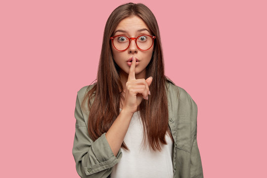 Shh, dont make noise! Scared young comely woman shows hush gesture, afraids of telling secret to someone, hopes for loyalty has fearful expression, wears spectacles, oversized shirt, poses indoor.