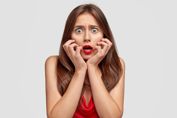 Wall Mural - Fearful nervous woman keeps hands on cheeks, stares with bugged eyes, opens mouth from fright, has red lips, poses against white background. Shocked stupefied wife notices husband with lover