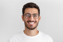 Daylight Portrait Of Young Handsome Caucasian Man Isolated On Grey Background, Dressed In White T-shirt And Round Eyeglasses, Looking At Camera And Smiling Positively