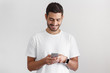 © Damir Khabirov - Daylight portrait of young european caucasian man isolated on gray background wearing white t-shirt standing in front of camera, looking attentively with smile at screen of smartphone he is holding