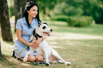 Young beautiful brunette cheerful girl in blue dress have fun and playing with her male white dog outdoor at nature. Nice looking woman loves kind pet. Happy best friends hugs. Domestiic animal smile.