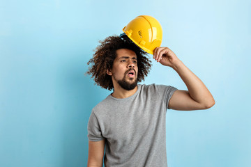 Wall Mural - A curly-headed handsome man wearing a gray T-shirt is standing with a safety helmet and looking sideways over the blue background.