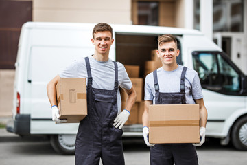 Wall Mural - Two young handsome smiling workers wearing uniforms are standing next to the van full of boxes holding boxes in their hands. House move, mover service.