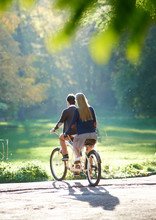 Back View Of Young Couple, Man And Blond Long-haired Woman Riding Tandem Double Bike Over The Edge Of Paved Path To Green Grassy Lawn On Blurred Dense Woody Forest Lit By Bright Sun Background.