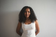 © Anatoliy Karlyuk - Fashionable young dark skinned woman wearing white tank top smiling and keeping hands behind her back standing isolated against blank studio wall background with copy space for your text