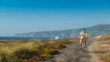 Mature Man (55-60) Cycles Along A Bumpy Asphalt Path Overlooking Praia Do Guincho And Roca Do Cabo Near Cascais, Portugal During A Windy Summer Day