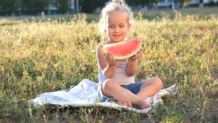 Wall Mural - A cute little blonde is sitting in the park on the grass and gaily eating a watermelon.