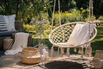 a beige string swing with a pillow on a patio. wicker baskets, a rug and a blanket on a wooden deck 