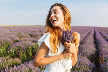 Canvas Print - Photo of smiling joyful woman in dress holding bouquet with flowers, while walking outdoor through lavender field in summer