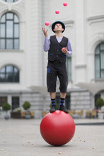 The Clown And Balancer Juggles With Pink Balls , Standing On A Big Red Ball In The Street Of A European City
