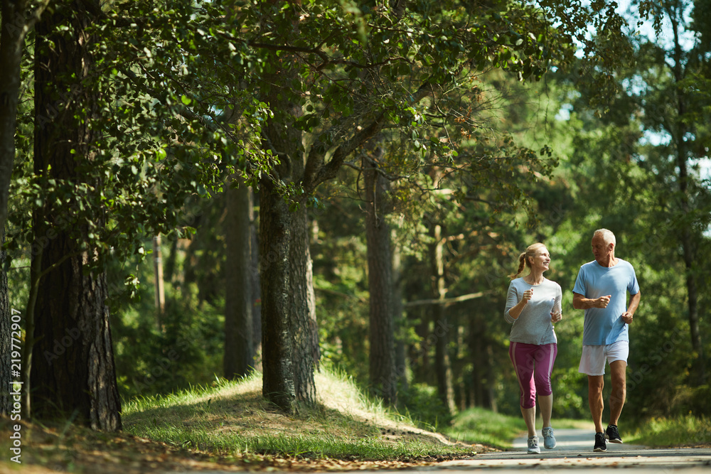 Healthy active senior couple in sportswear running among green trees on sunny day in summer - obrazy, fototapety, plakaty 