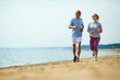 Active senior man and woman running down sandy beach with waterside on background