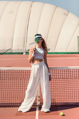 Wall Mural - stylish woman in white clothing and cap with tennis racket posing at tennis net on court