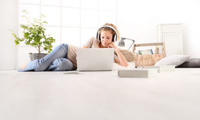 young woman with computer, headphones, smartphone and books, lying on the floor in living room on white wide window in the background