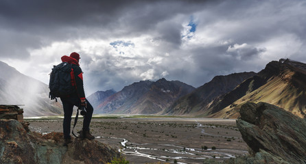 a man with backpack holding camera standing on cliff with mountains view and sunlight through cloud.