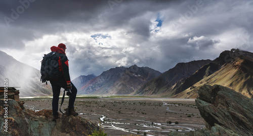 Fotomural a man with backpack holding camera standing on cliff with mountains view and sunlight through cloud