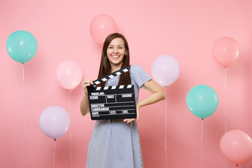 Portrait of smiling pretty young woman wearing blue dress holding classic black film making clapperboard on pink background with colorful air baloons. Birthday holiday party, people sincere emotions.