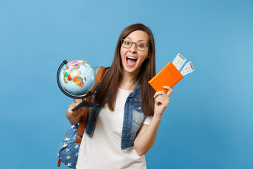 Young happy woman student in glasses with backpack holding world glove, passport, boarding pass tickets isolated on blue background. Education in university college abroad. Air travel flight concept.