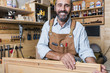 © tiero - smiling carpenter with long hair and beard wears leather apron inside his workshop. serene and satisfied expression. manual and traditional work.