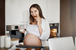 © Liubov Levytska - Pregnant woman do not share food. Charming and caring european wife with cute belly, standing near kitchen table with cookies on dripping-pan, biting cookies joyfully and holding glass of milk