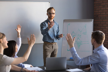 Workers raising hands asking questions during male speaker or coach flipchart presentation at office team training, people answering during corporate educational seminar, brainstorming at meeting