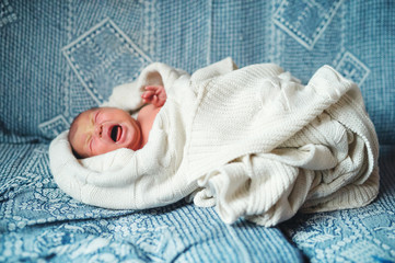 Wall Mural - Crying newborn baby lying on a sofa, covered by a white blanket. Side view.